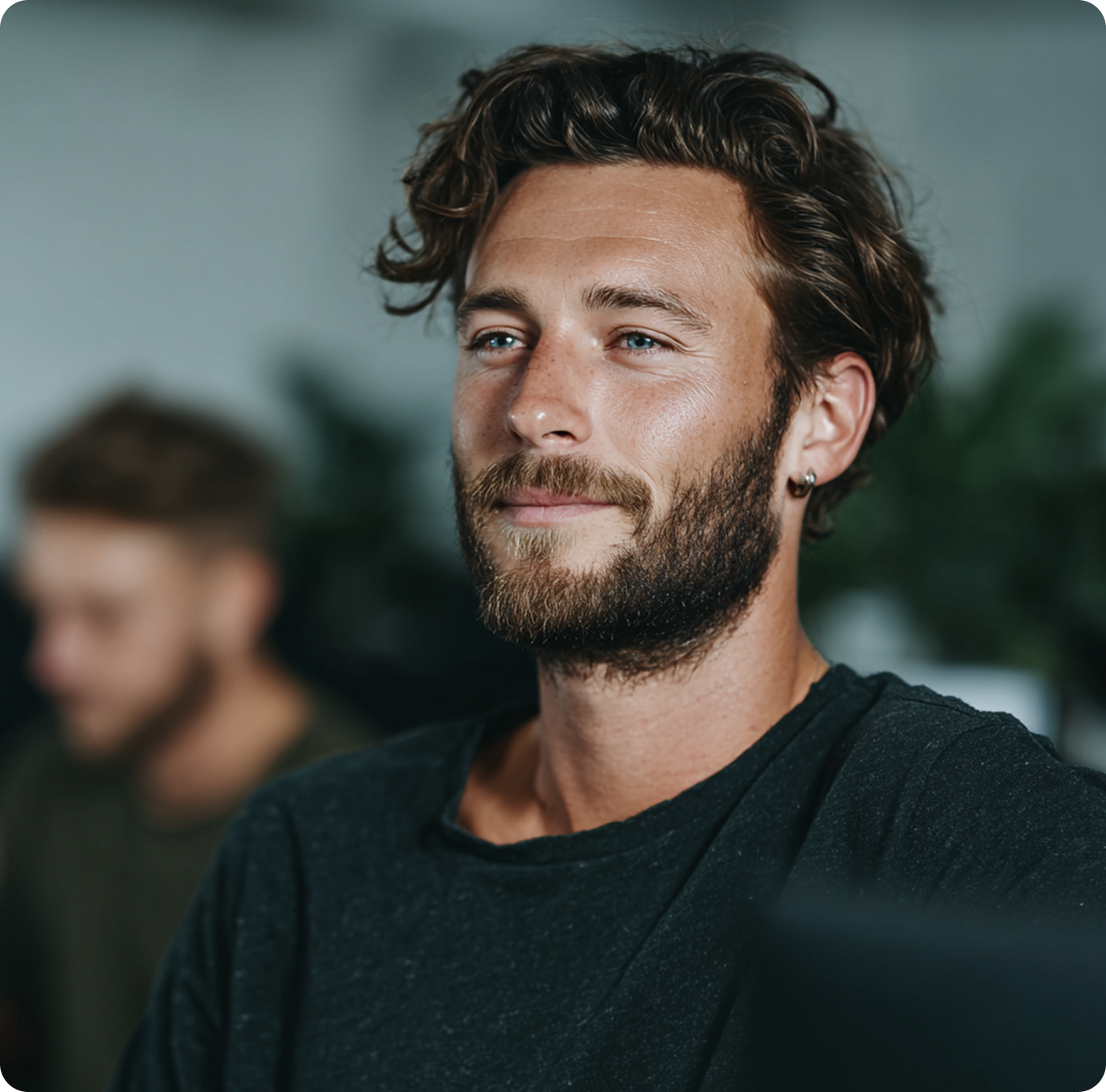 Bearded young man with wavy hair and earring wearing a dark shirt, with a blurred person in the background.