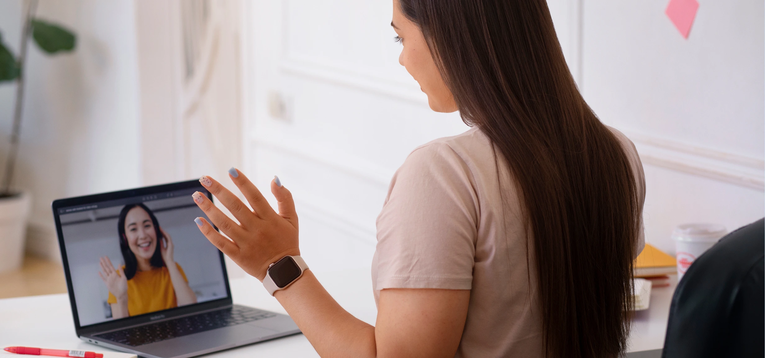 Woman with long brown hair waving during a video call with another smiling woman on a laptop screen.