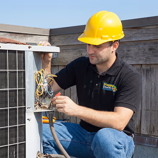 Technician wearing a yellow hard hat and black shirt repairing an outdoor HVAC unit with exposed wiring.