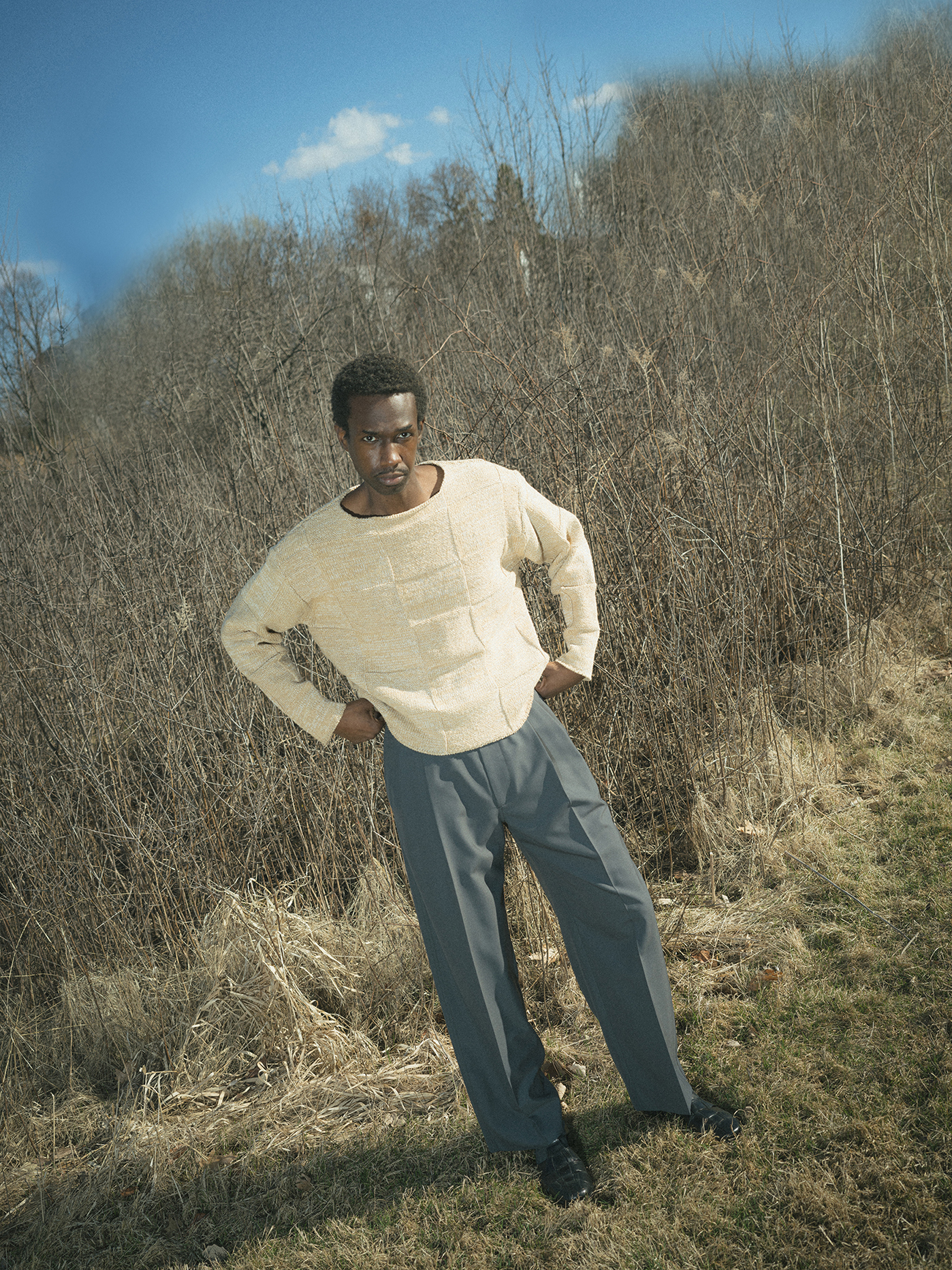 Man wearing AURALEE in cream sweater poses confidently in barren forest landscape under blue sky 