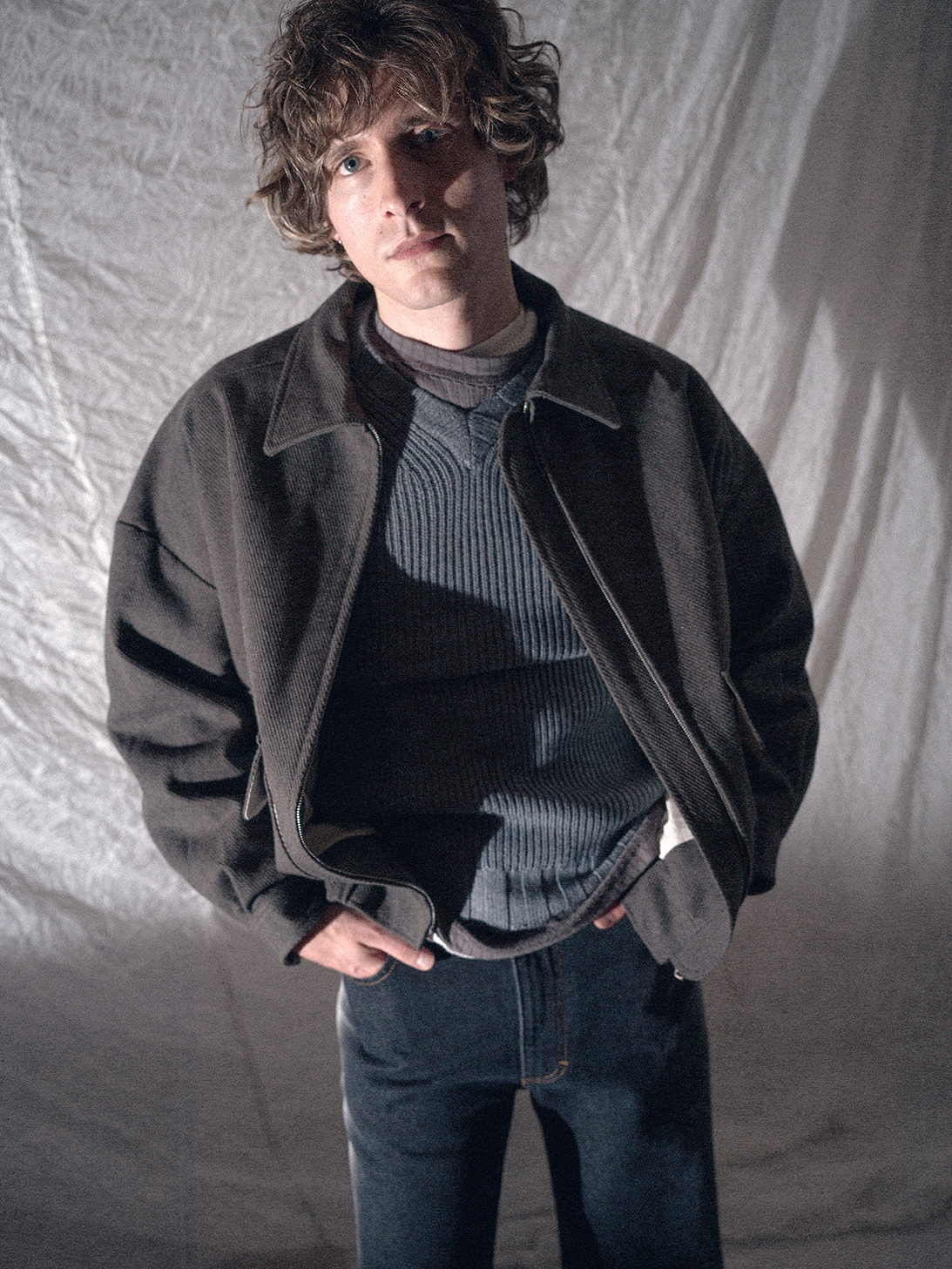 Portrait of a man in a dark jacket and textured sweater against a light draped backdrop