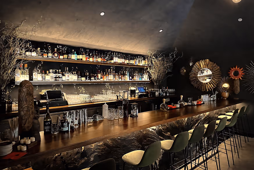 Stylish dimly lit bar with wooden counter, high green chairs, shelves stocked with bottles, and decorative mirrors on dark walls.
