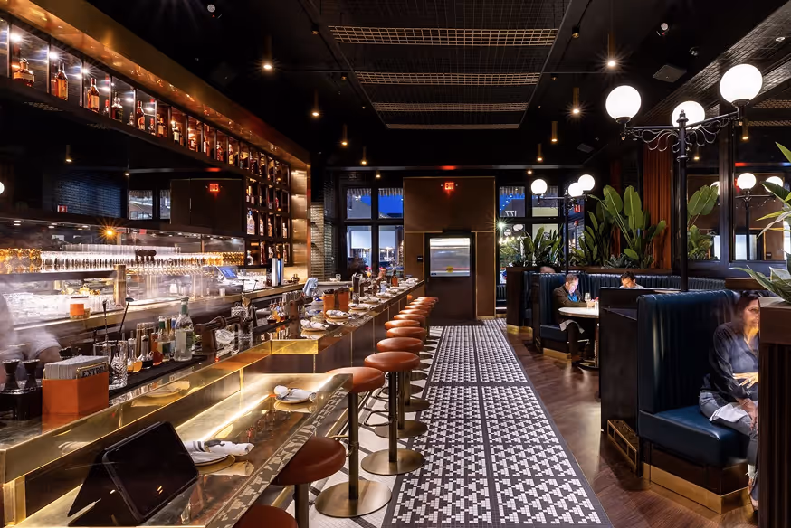 Modern bar interior with a long counter lined with red cushioned stools, patterned floor walkway, and booth seating with plants and globe lights.