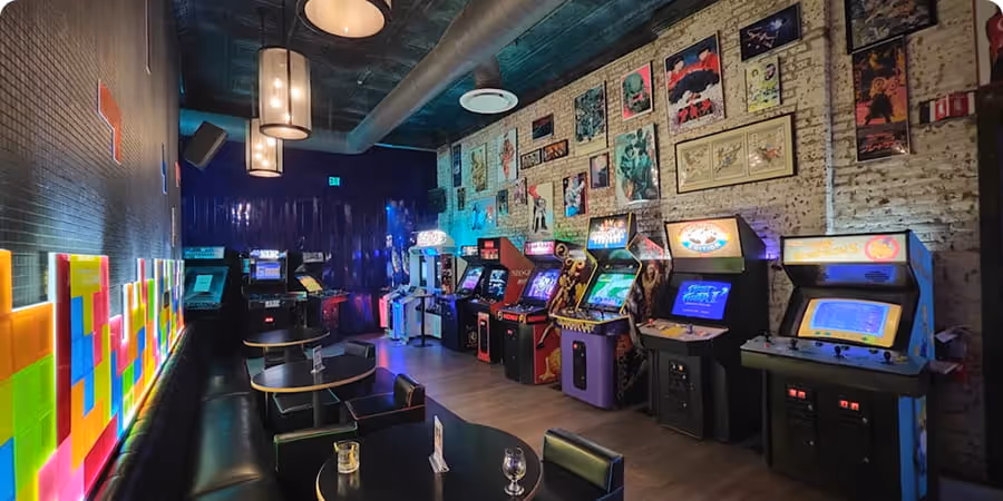 Interior of an arcade bar with rows of illuminated vintage arcade game machines against a brick wall decorated with posters and art, and seating with tables and chairs nearby.