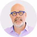 Bald man with glasses and beard wearing a light purple shirt smiling against a light background.