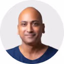 Smiling bald man wearing a dark blue shirt against a light gray background.