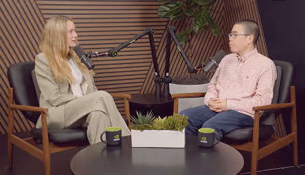 Two people sitting in chairs facing each other, speaking into microphones, with a small table holding plants and two mugs between them.