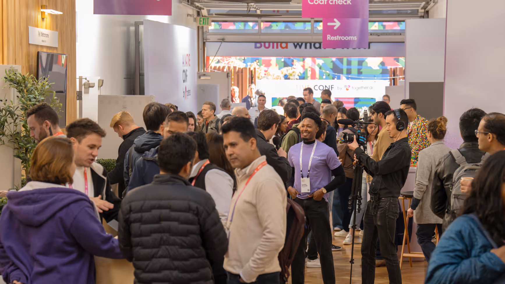 Crowd of diverse people in a busy conference hallway with a cameraman filming and signs indicating coat check and restrooms.