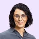Woman with short curly dark hair and glasses wearing a gray collared shirt on light purple background.