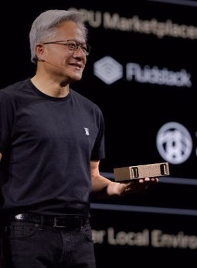 Man wearing glasses and black shirt holding a rectangular device on stage with presentation screen behind.