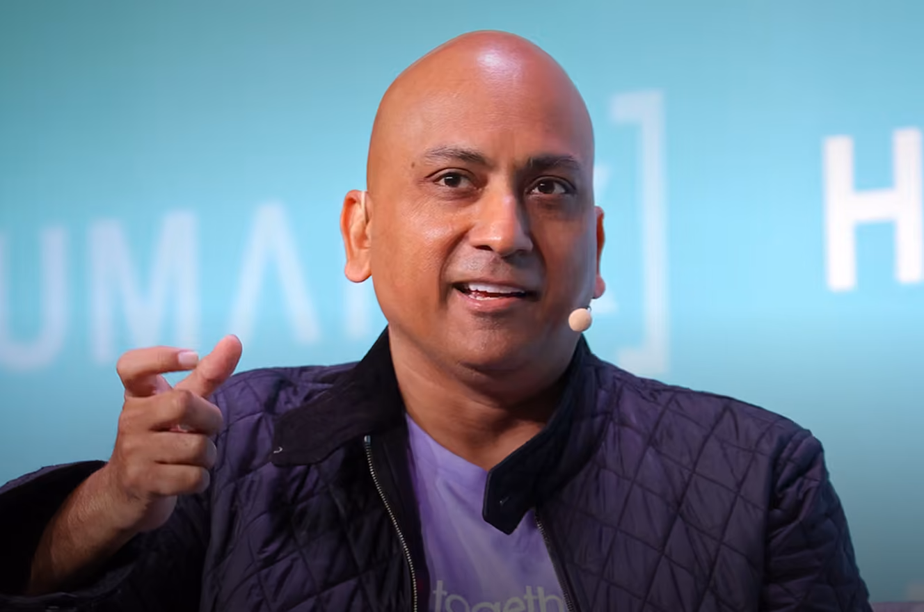 Bald man speaking with a headset microphone against a blue background with blurred white text.