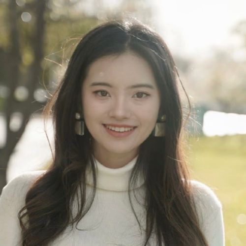 Young woman with long dark hair smiling outdoors wearing a white turtleneck and statement earrings.