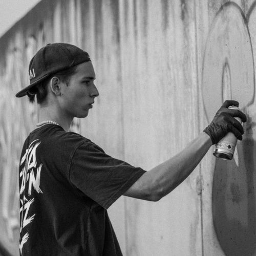 Young man wearing a cap sprays graffiti on a wall with a spray paint can in black and white.