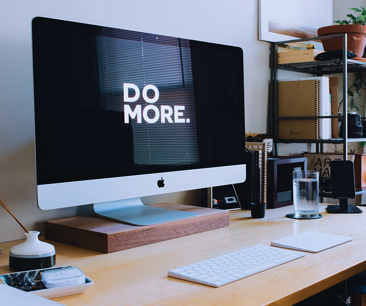 Modern workspace with an iMac displaying 'DO MORE.' on screen, keyboard, glass of water, and organized shelves.