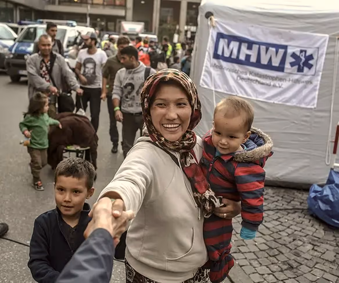 Smiling woman in a headscarf holding a baby and shaking a person's hand on a crowded street with people and a tent in the background.