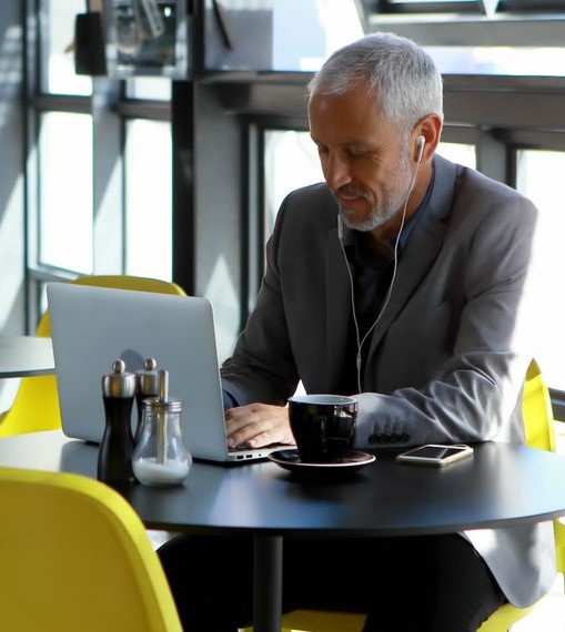 Man in a gray suit sitting at a round table working on a laptop with earphones, coffee cup, and smartphone nearby in a bright café.