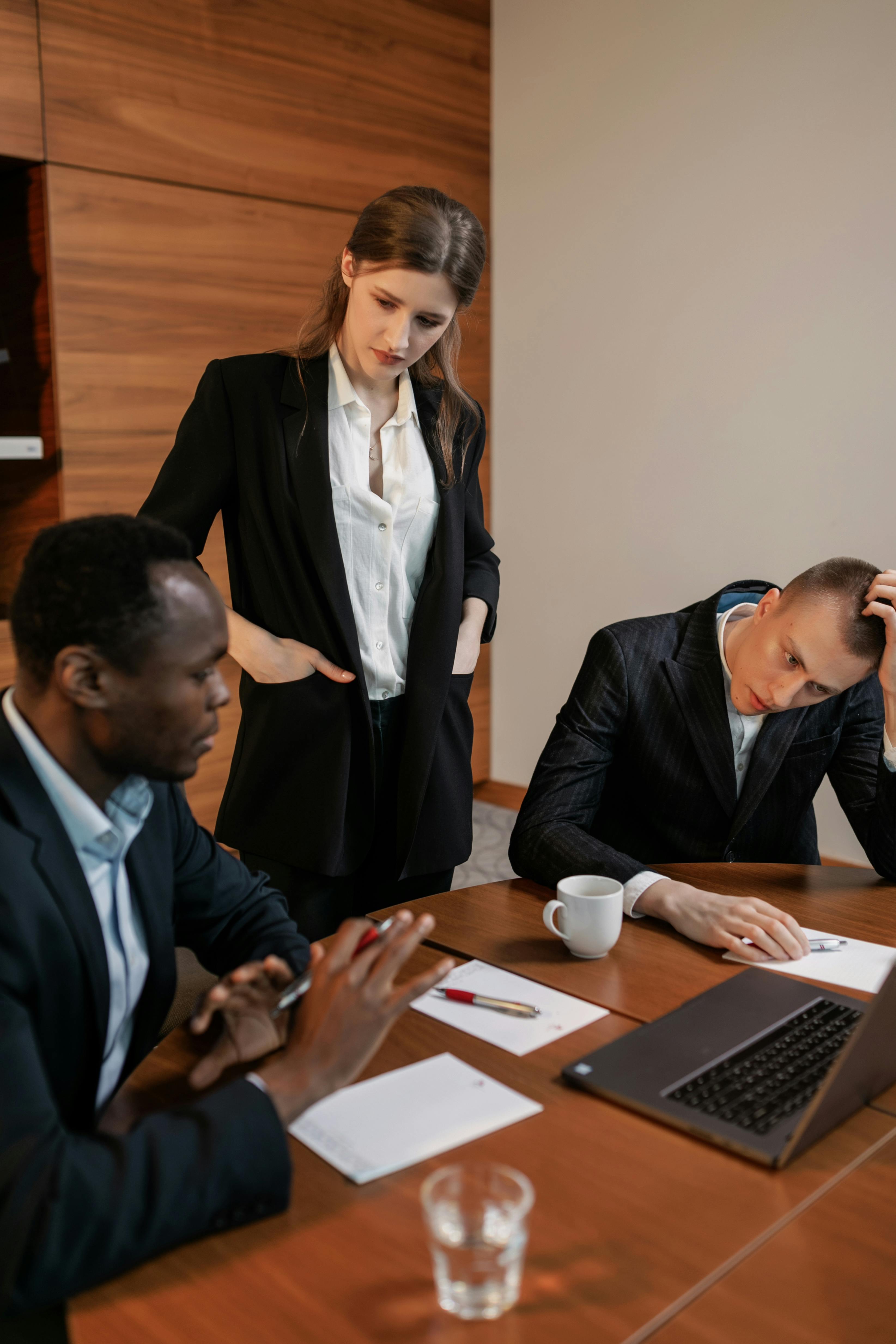 Three business professionals in a meeting room, two men seated with notebooks and a laptop, and a woman standing and looking at the laptop.