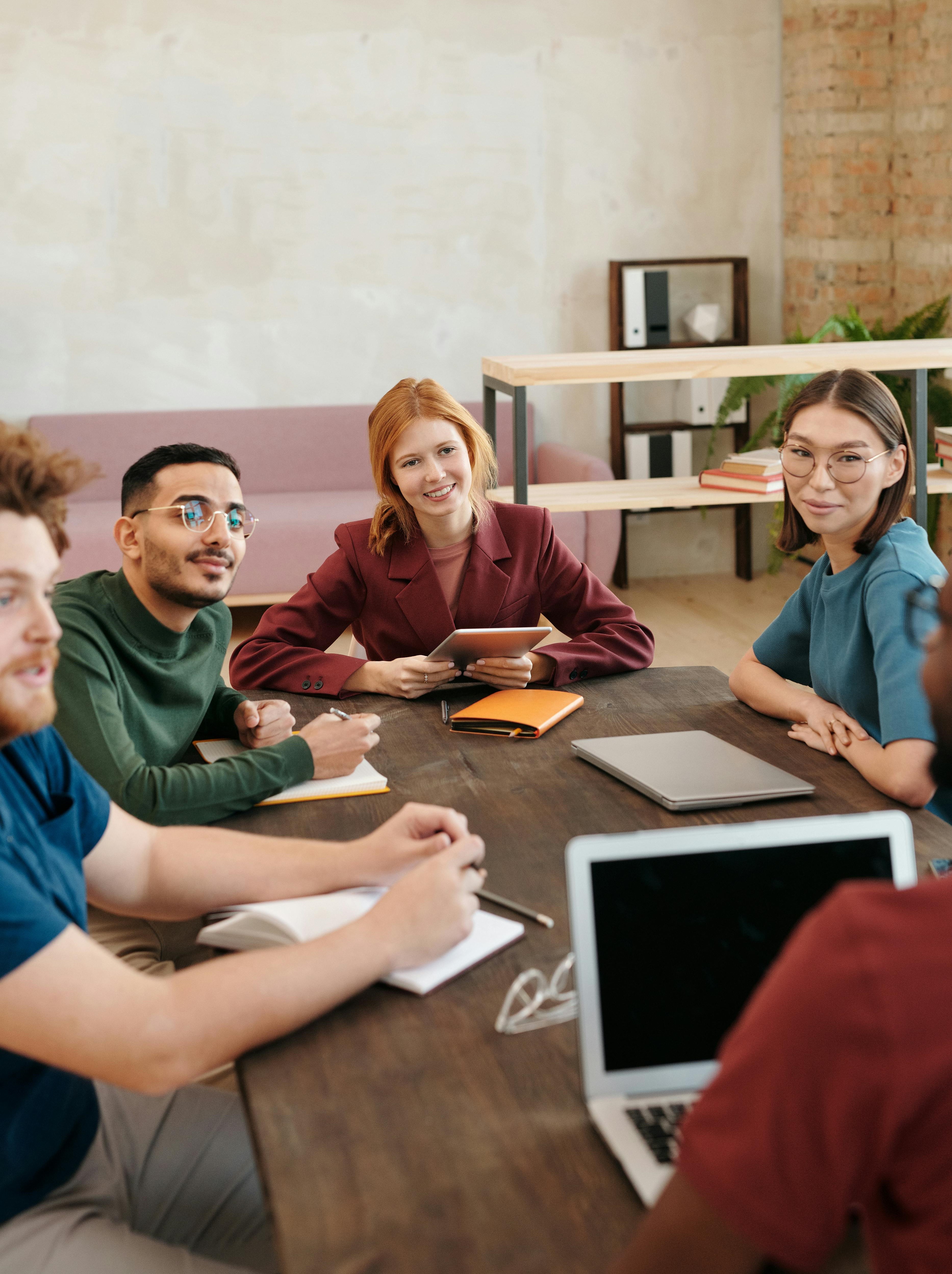 Four diverse young adults sitting around a wooden table in a casual modern workspace, engaged in a discussion with notebooks, a tablet, and a laptop.