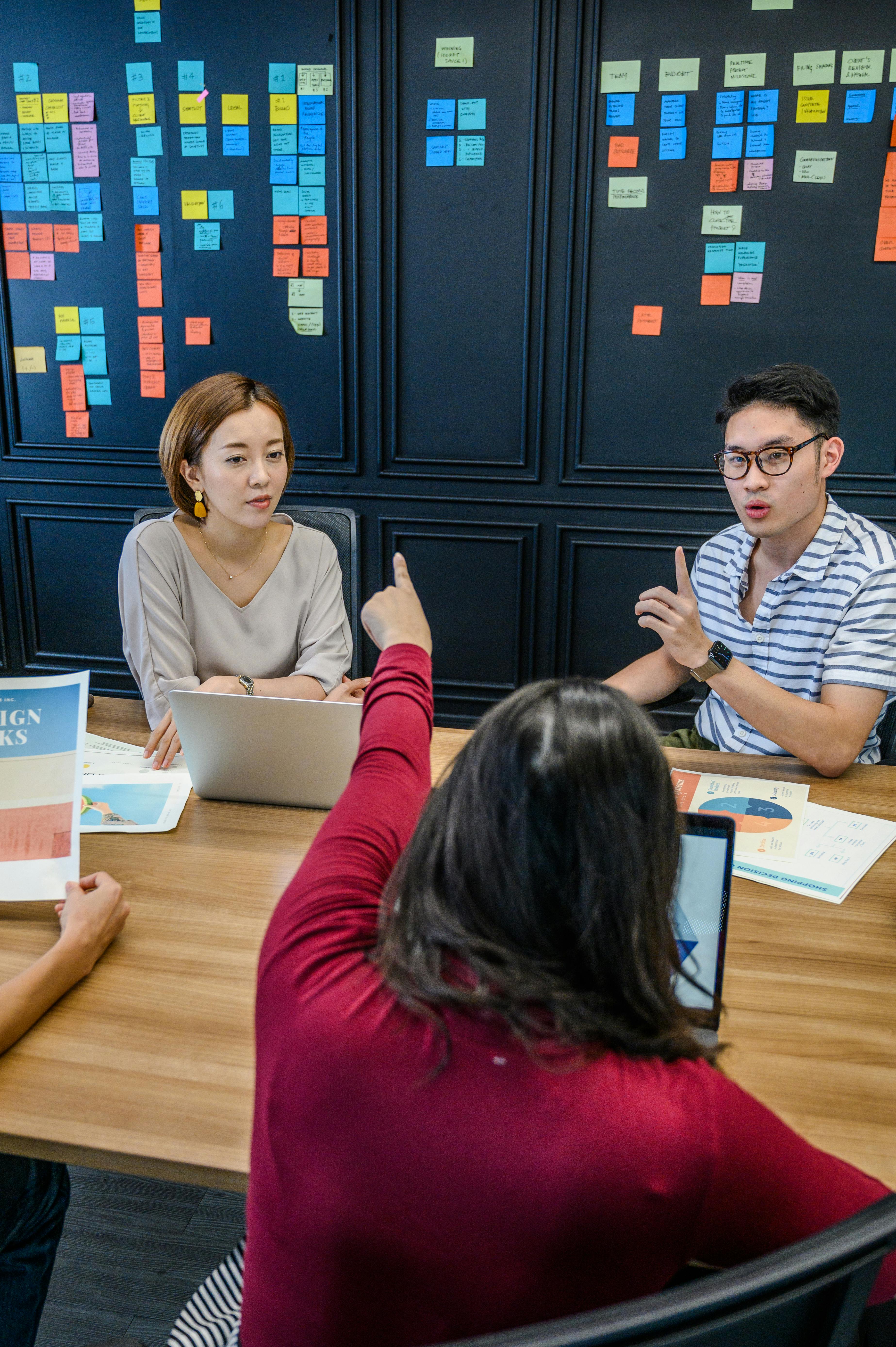 Three people engaged in a discussion at a wooden table with laptops and printed charts, colorful sticky notes on black wall in background.