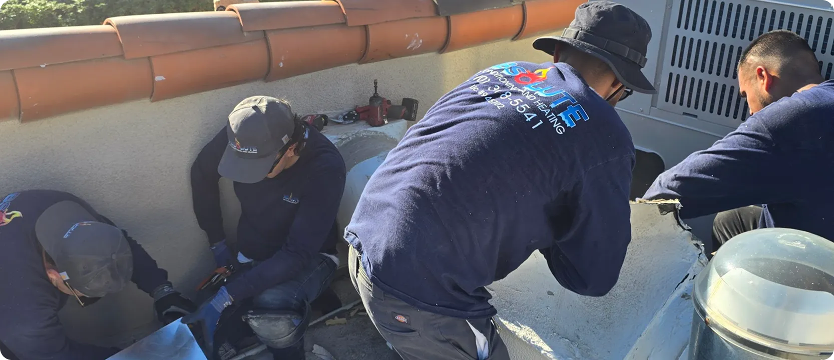 Four workers in blue uniforms repairing or installing HVAC equipment outdoors near a rooftop wall.