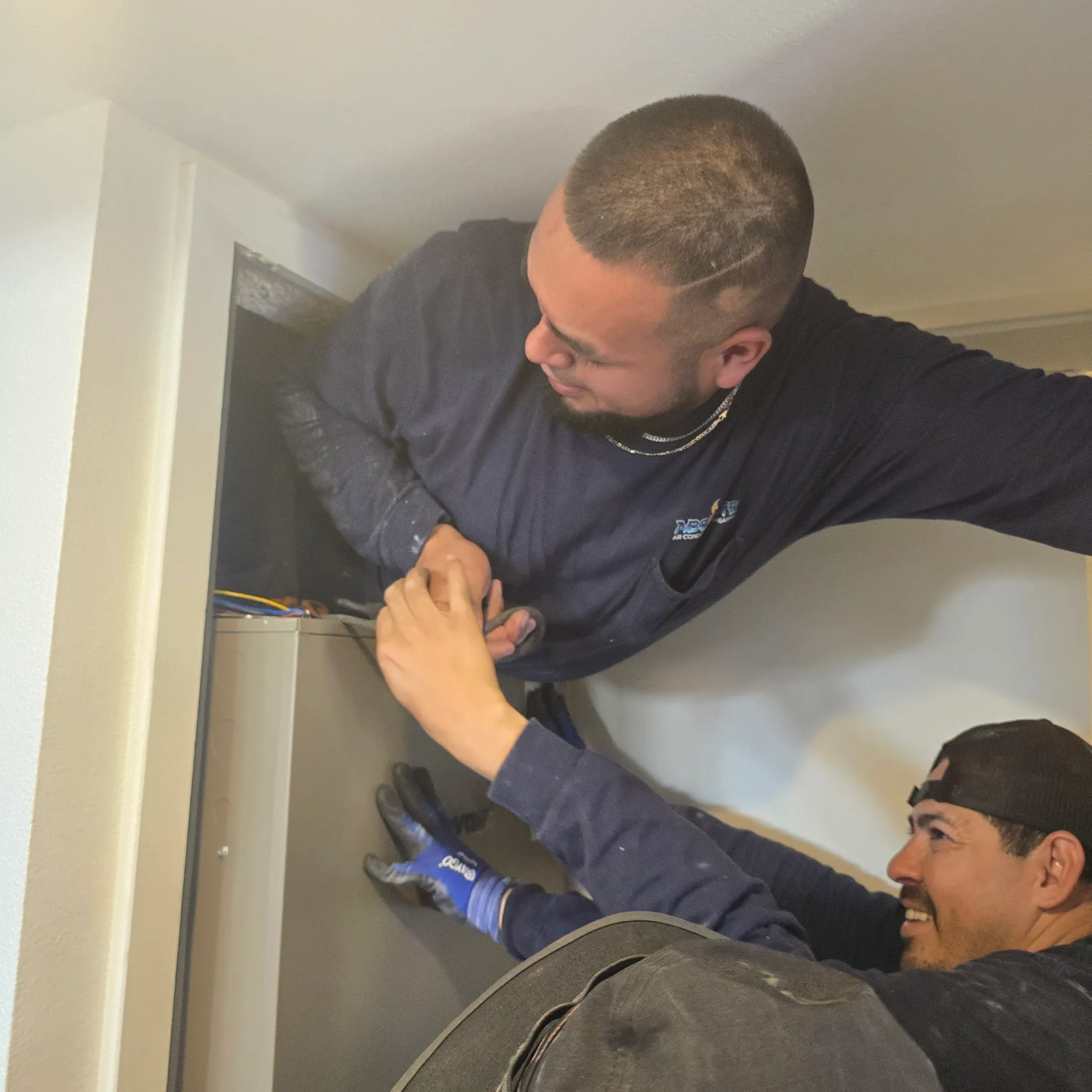 Two men working together to install or repair an HVAC unit inside a wall cavity.