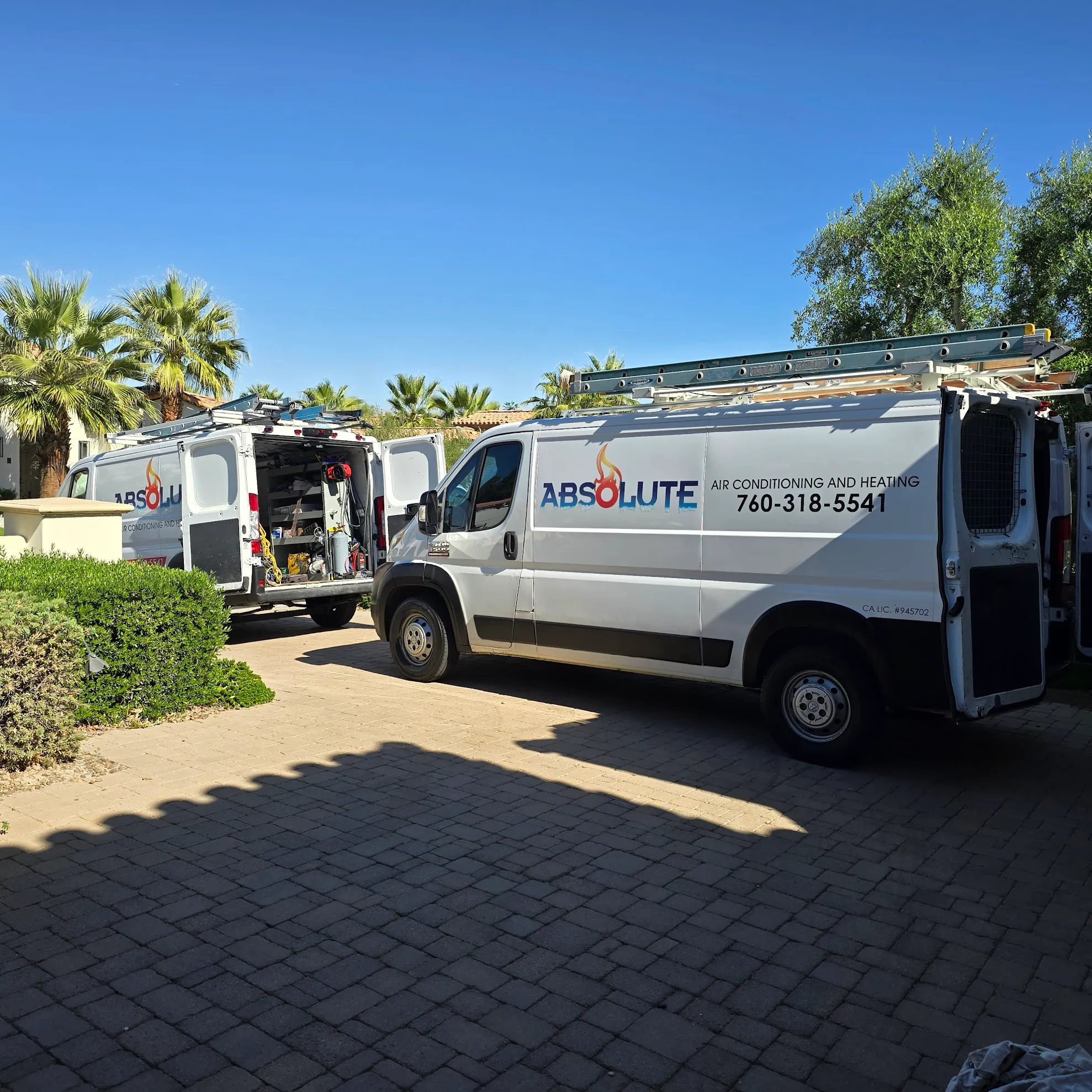 Two white Absolute Air Conditioning and Heating vans parked on a driveway with ladders on their roofs and rear doors open on one van showing equipment inside.