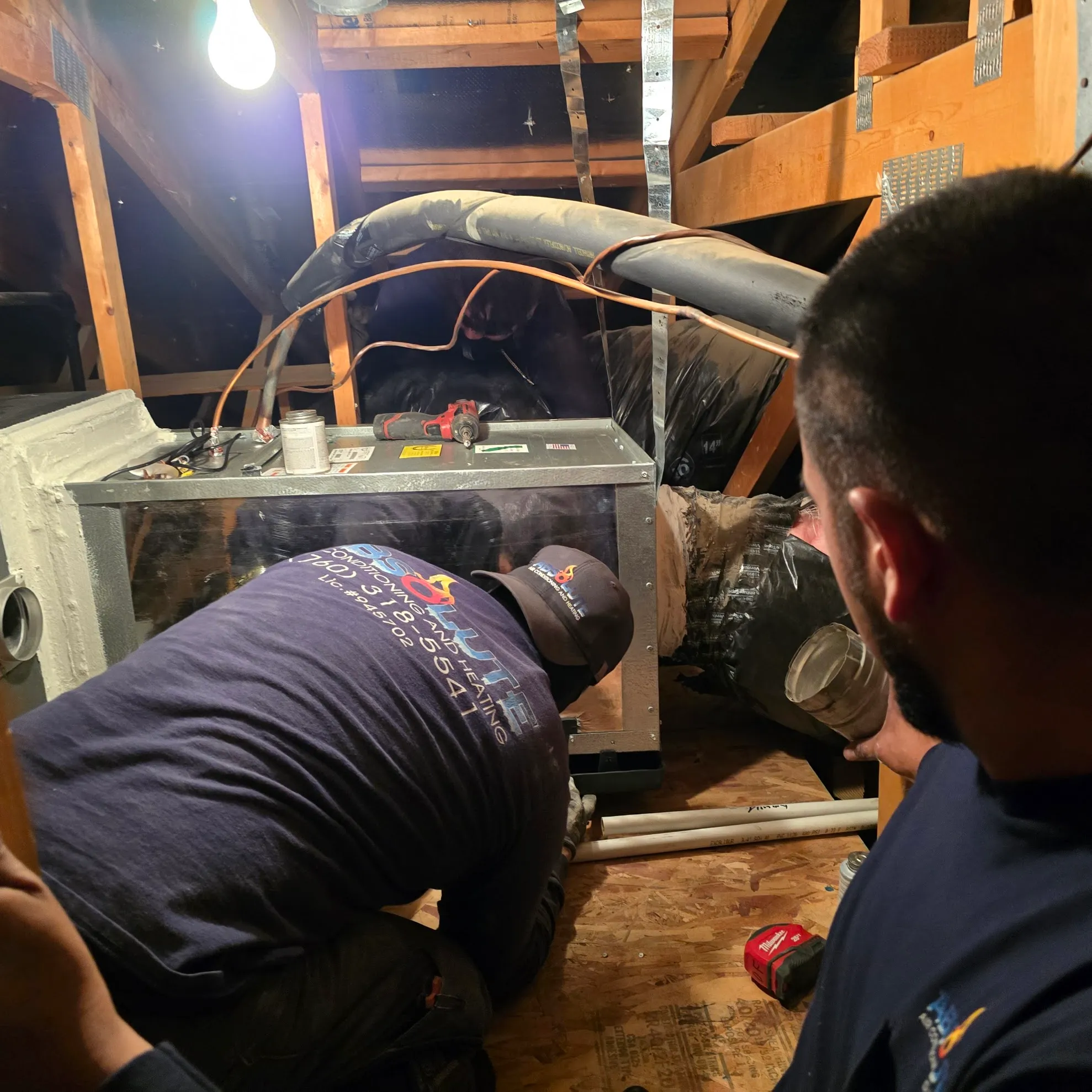 Two HVAC technicians installing or servicing equipment in an attic space with ductwork and tools around them.