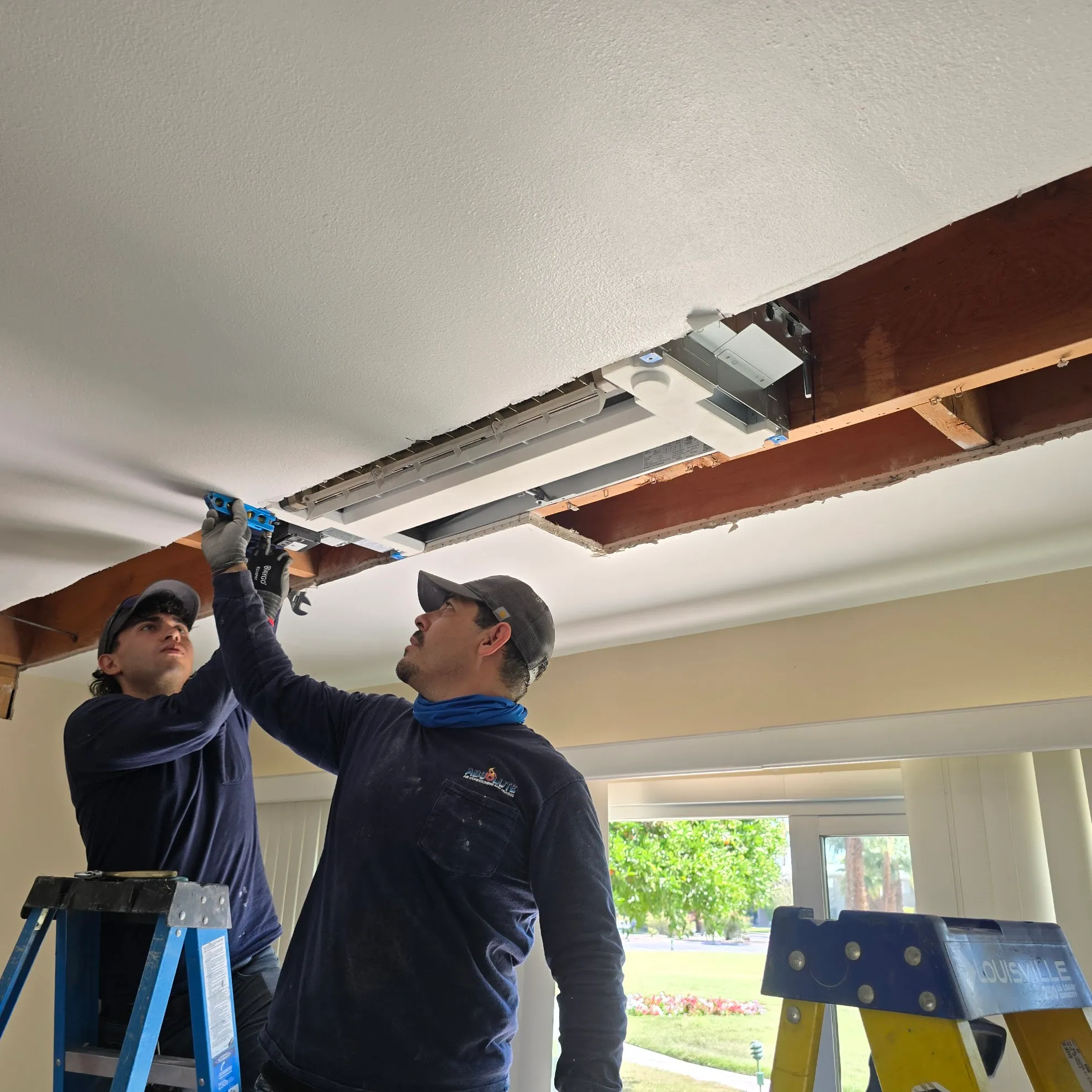 Two workers installing an air conditioning unit inside a partially opened ceiling, one standing on a blue ladder, the other reaching up.