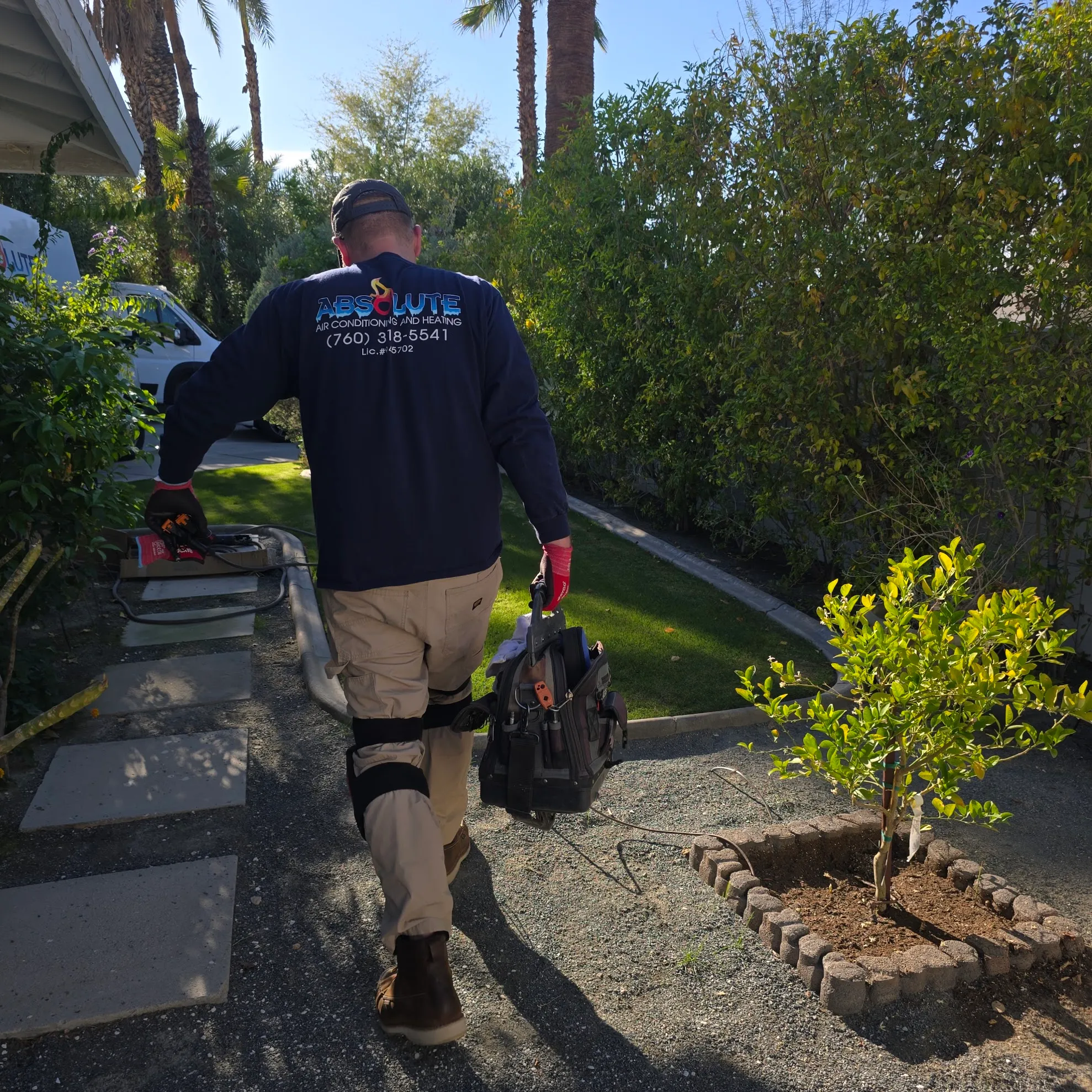Technician walking on a garden path carrying a toolbox and wearing an Absolute Air Conditioning and Heating shirt.