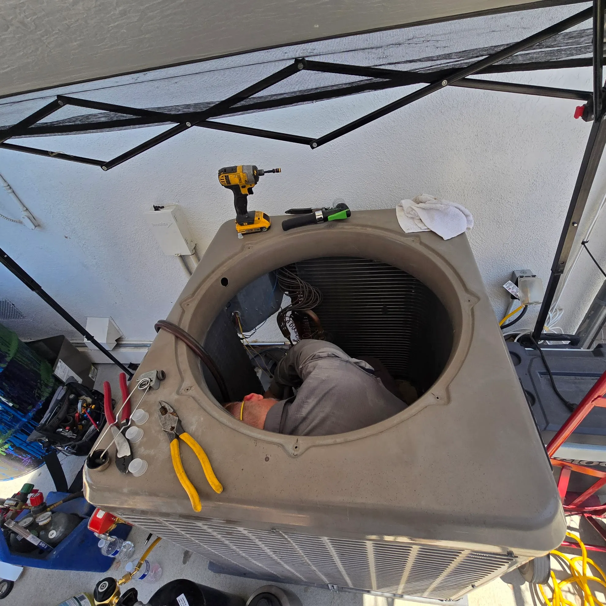 Technician working inside an open outdoor air conditioning unit with tools placed on top and nearby equipment.