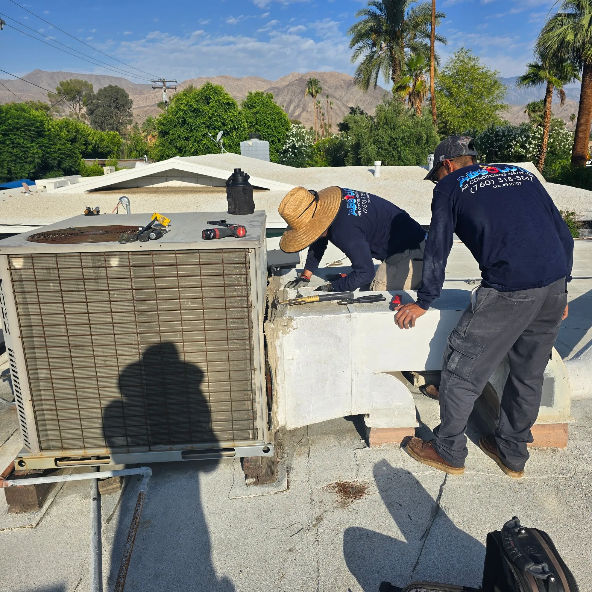 Two technicians in blue shirts working on an air conditioning unit on a rooftop with tools nearby and mountains in the background.