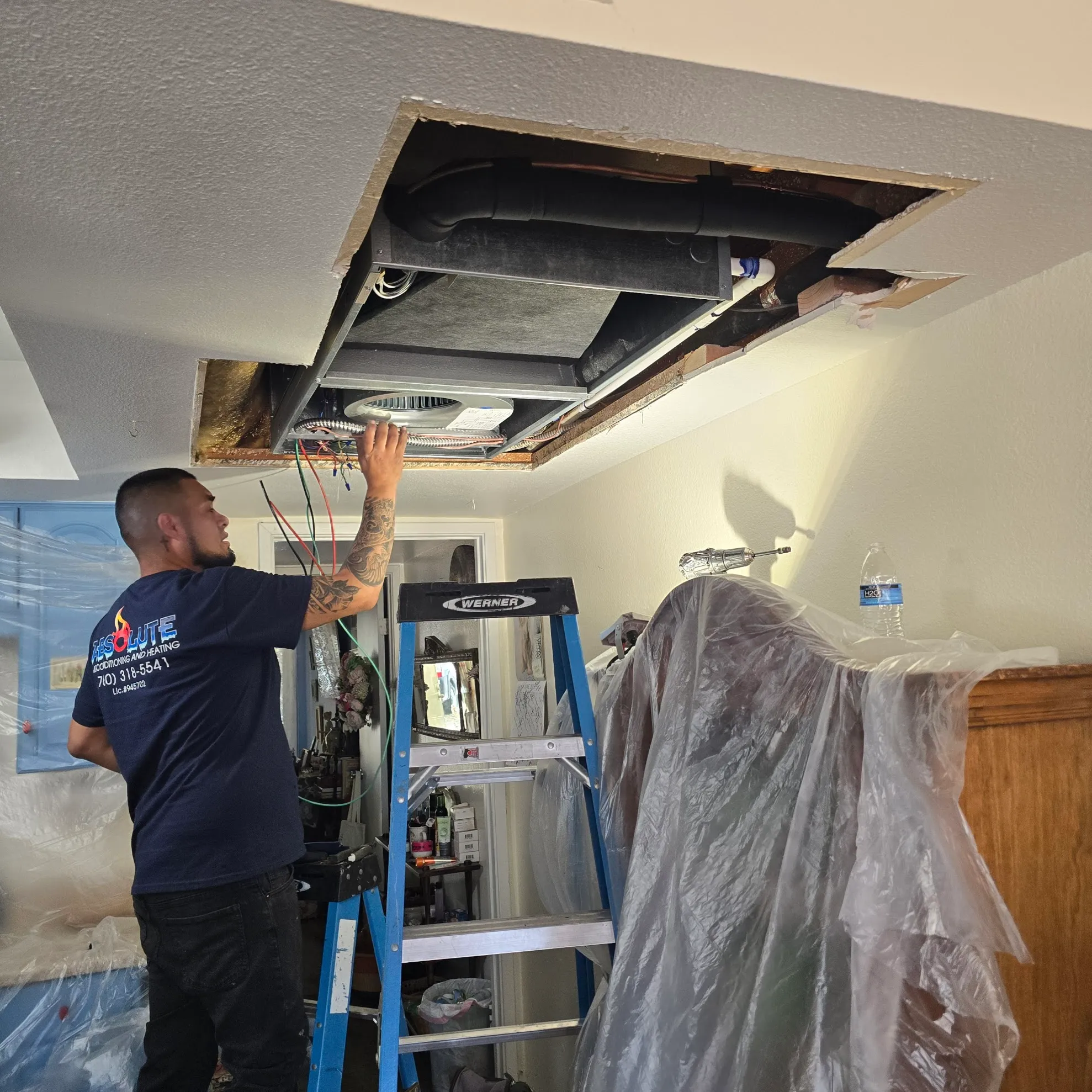 Technician installing or repairing an HVAC system in a ceiling, standing on a ladder with covered furniture nearby.