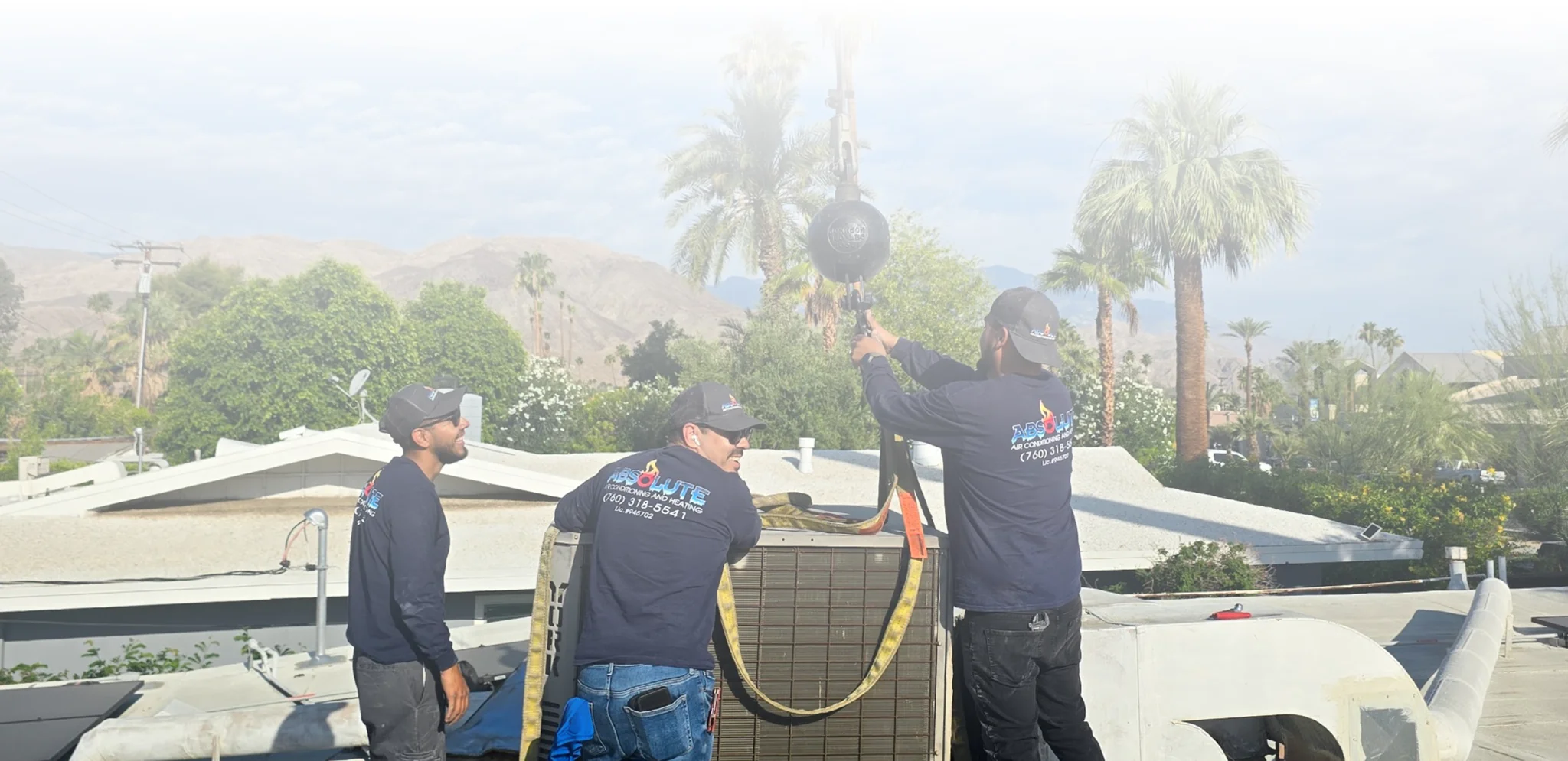Three workers in dark shirts with 'Absolute Air Conditioning and Heating' logos performing maintenance on rooftop HVAC equipment surrounded by palm trees and mountains.