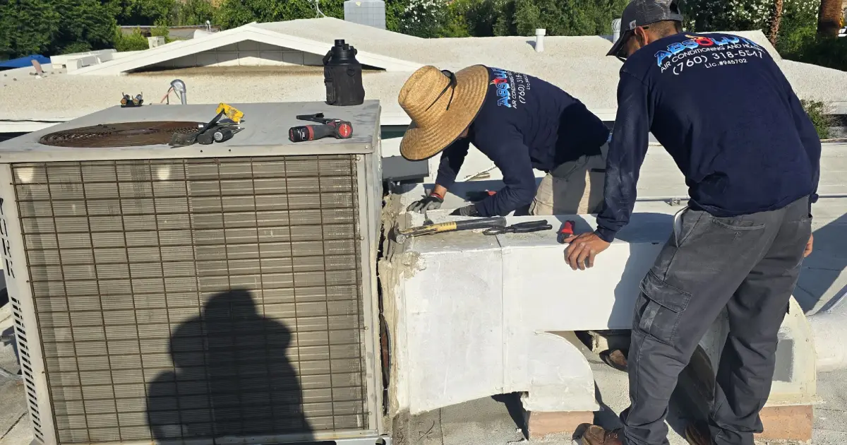 Two HVAC technicians wearing uniforms working on an air conditioning unit on a rooftop with tools placed on top.