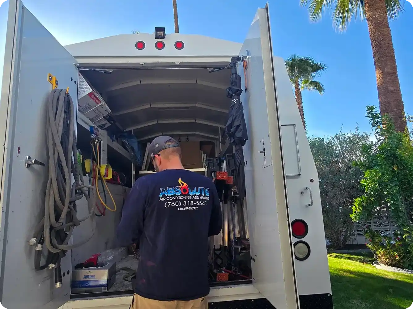 Technician wearing an Absolute Air Conditioning and Heating shirt standing at the open back of a service van with tools inside, outdoors with palm trees and greenery.