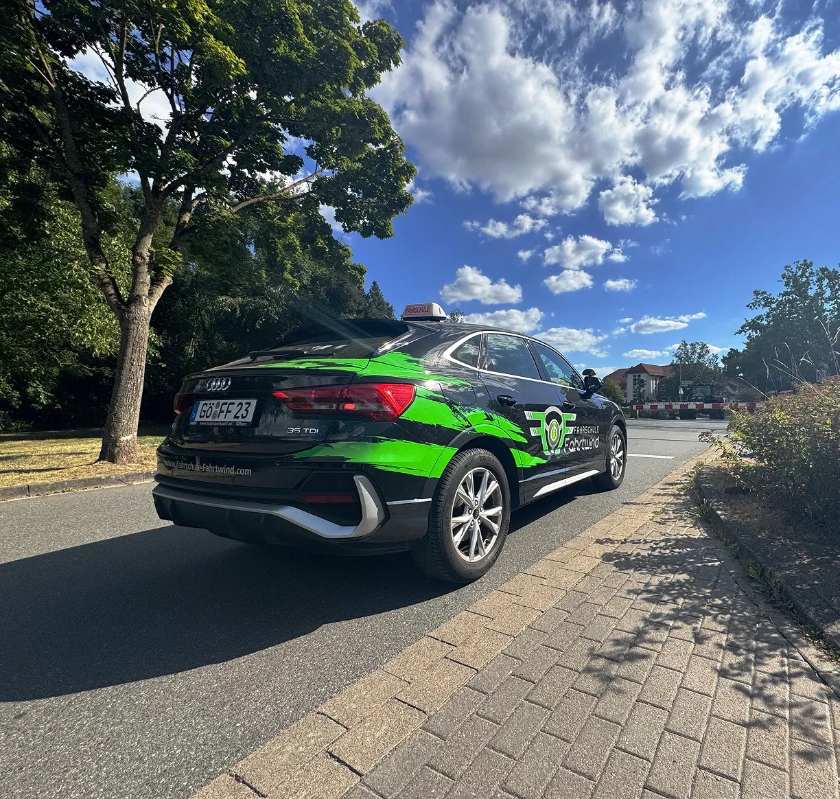 Schwarzes Audi-Fahrschulauto mit grünen Akzenten und Fahrterschule-Fahrwind-Logo steht auf Straßenrand unter blauem Himmel mit Wolken.