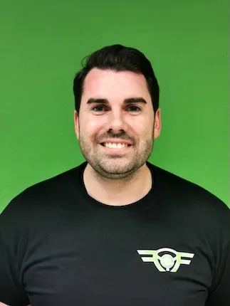 Lächelnder Mann mit dunklen Haaren und Bart vor grünem Hintergrund, trägt schwarzes T-Shirt mit Logo.