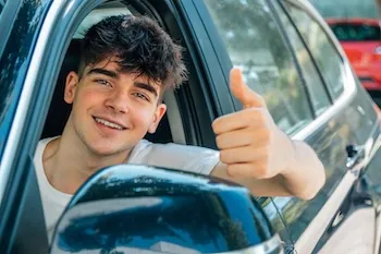 Junger Mann sitzt im Auto und zeigt Daumen hoch aus dem Fenster.