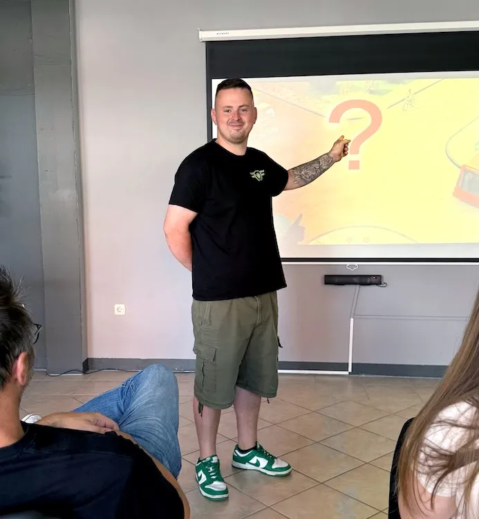 Ein Mann in schwarzem T-Shirt und grünen Shorts zeigt auf ein großes Fragezeichen auf einer Projektionswand, während andere Personen ihm zuhören.