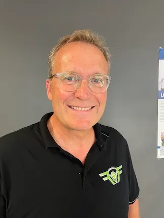 Älterer Mann mit grauen Haaren und Brille, lächelnd, trägt ein schwarzes Polohemd mit grünem Logo vor grauer Wand.