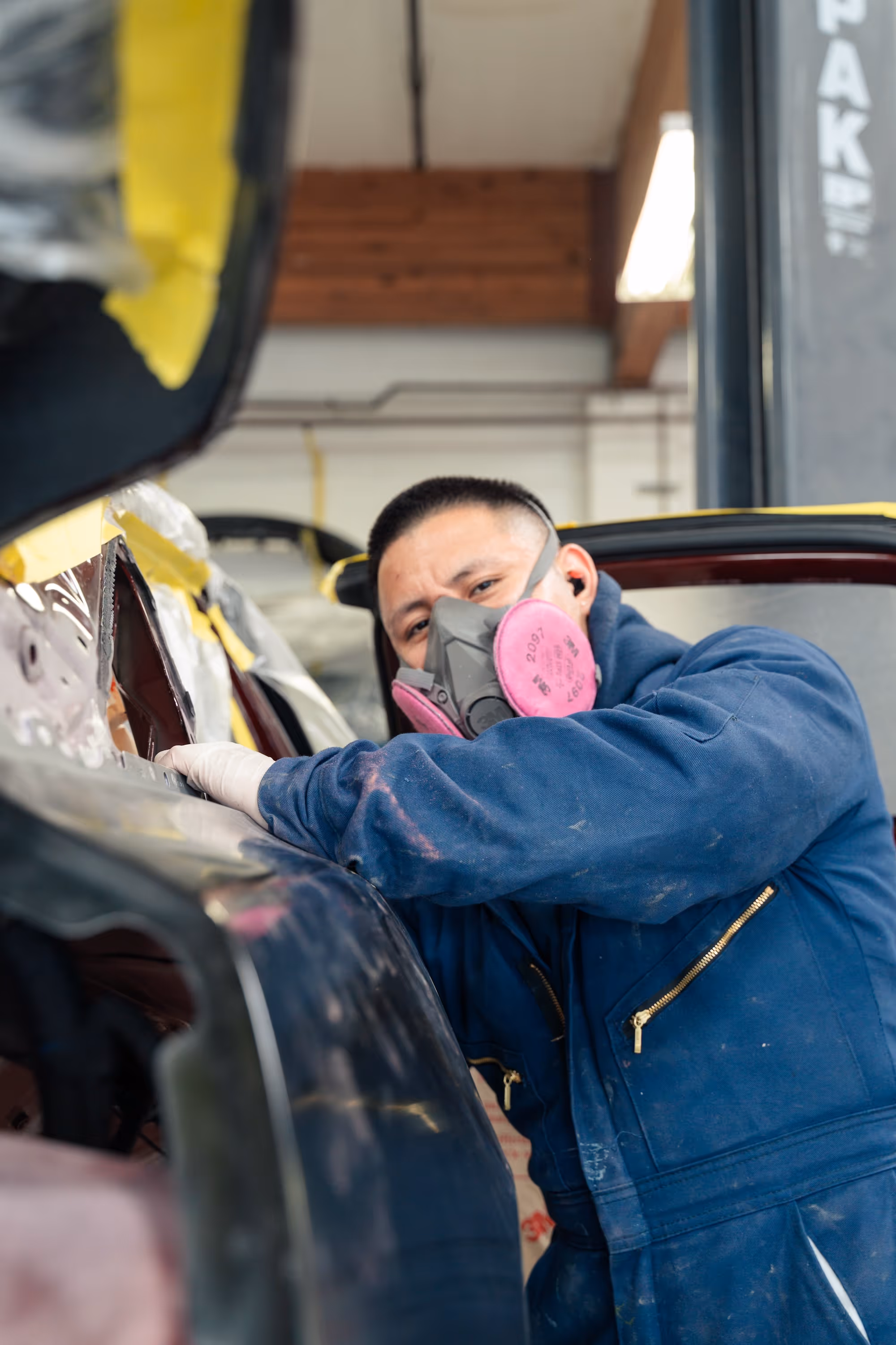 Collision repair expert wearing a protective mask leaning against the body of a car.
