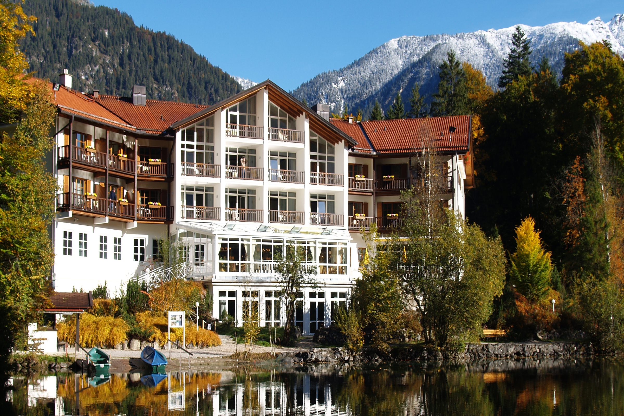 Hotelgebäude mit mehreren Balkonen am Ufer eines ruhigen Sees, im Hintergrund bewaldete Berge mit schneebedeckten Gipfeln.