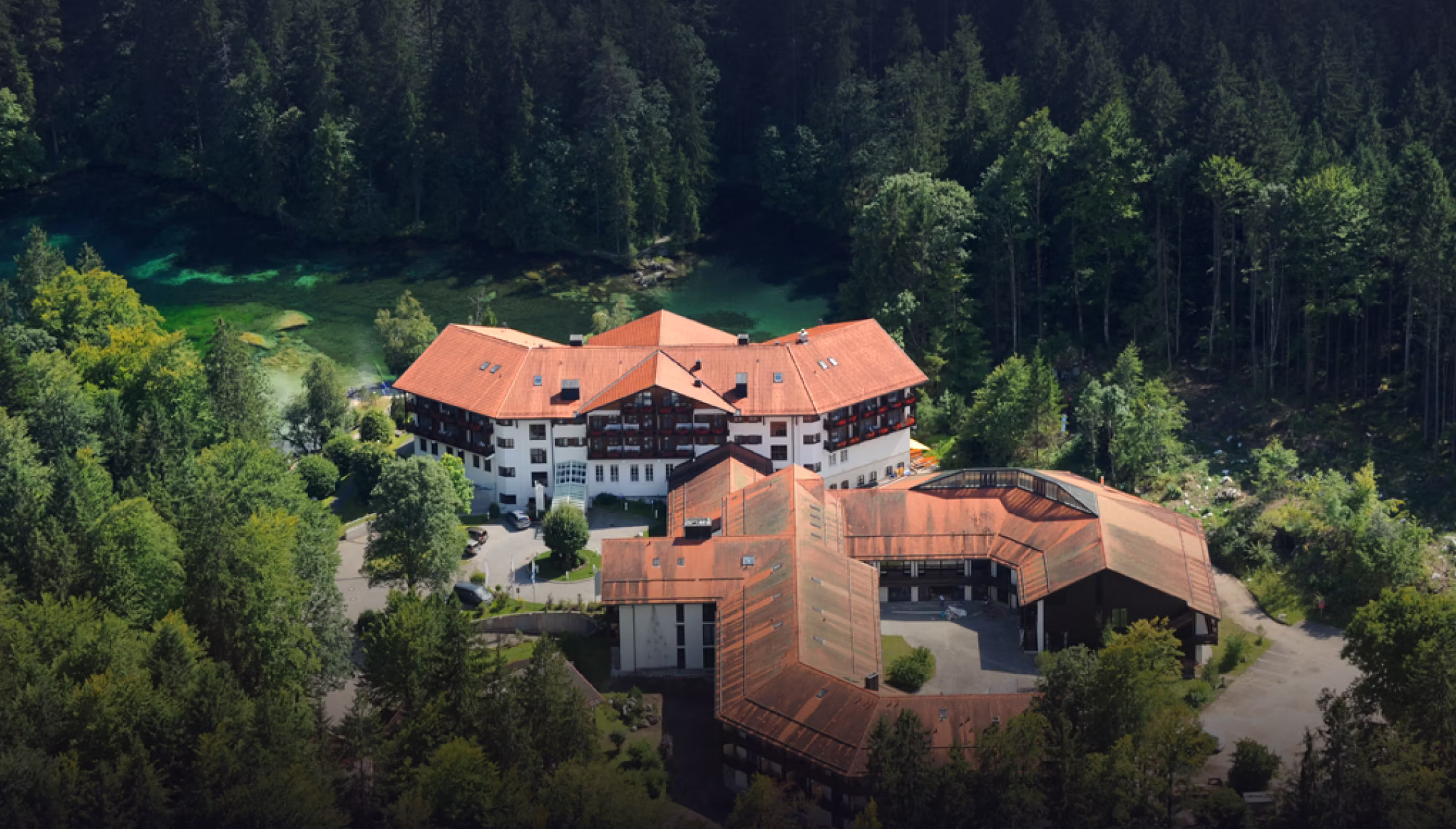 Luftaufnahme eines großen Gebäudekomplexes mit roten Dächern, umgeben von dichter grüner Waldlandschaft und einem kleinen See.