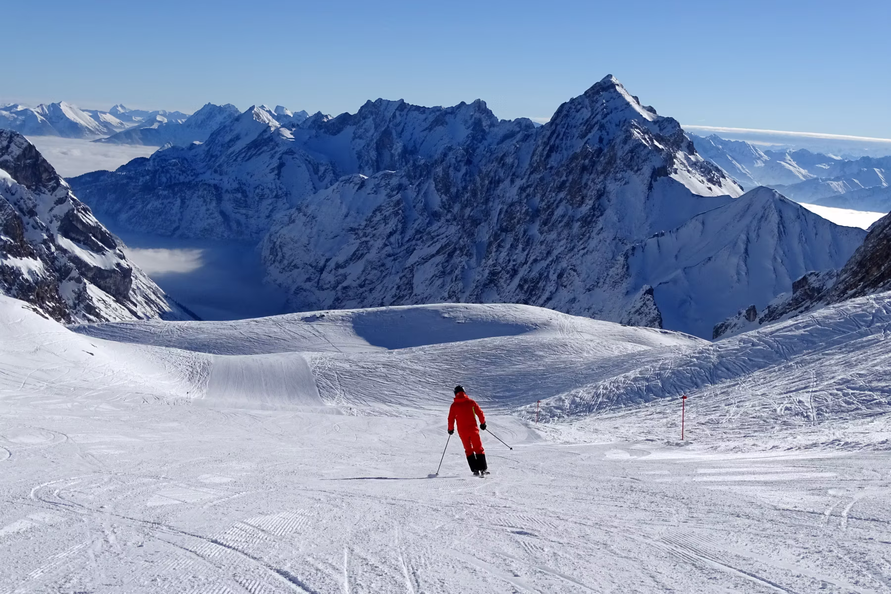 Skifahrer in rotem Anzug fährt auf einer schneebedeckten Piste vor schneebedeckten Berggipfeln unter klarem Himmel.