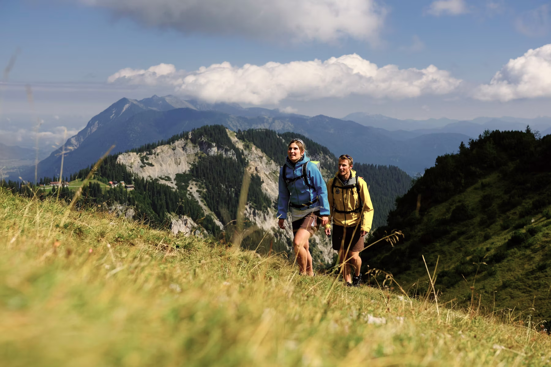 Zwei Wanderer mit Rucksäcken steigen durch eine grüne Berglandschaft mit bewaldeten Hügeln und bewölktem Himmel.