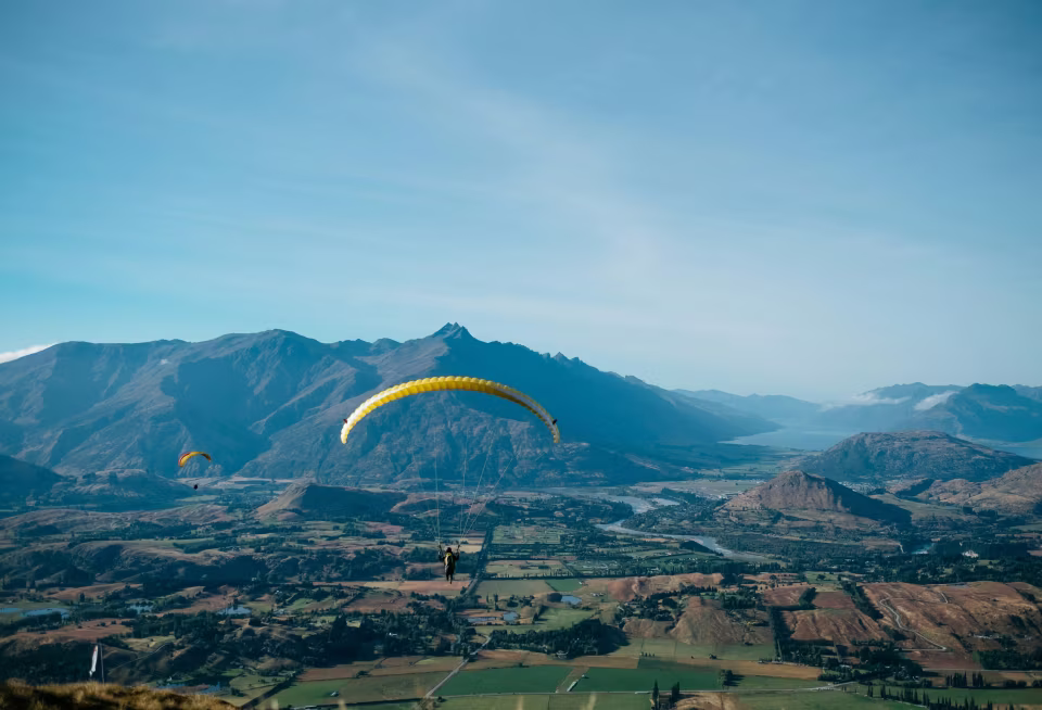 Zwei Gleitschirmflieger über grüner Landschaft mit Bergen unter blauem Himmel.