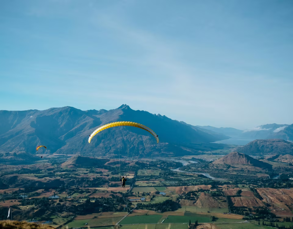 Zwei Gleitschirmflieger schweben über ein Tal mit grünen Feldern vor bewaldeten Bergen unter einem blauen Himmel.