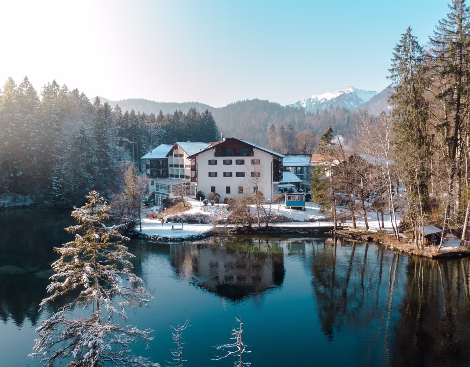 Ein weißes Gästehaus an einem See mit Schnee bedeckten Bäumen und Bergen im Hintergrund an einem klaren Wintertag.