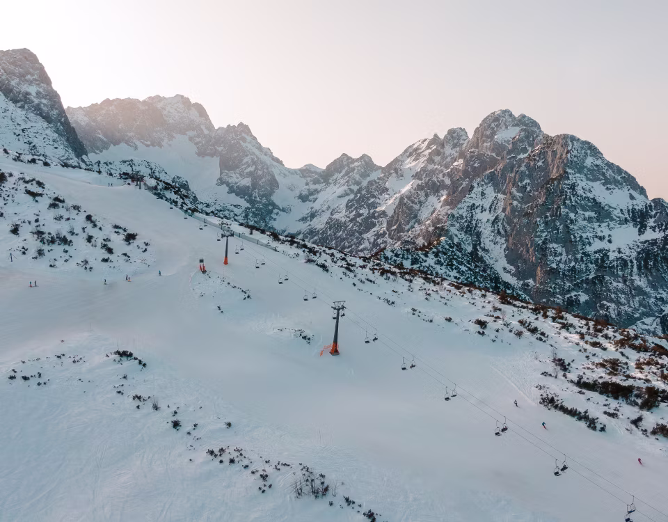 Verschneite Skihänge mit Sessellift vor hohen, felsigen Bergen bei Sonnenuntergang.