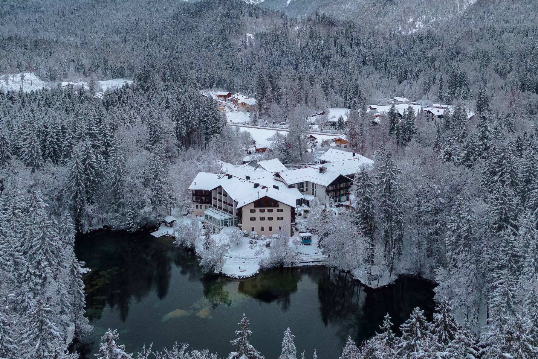 Verschneites Dorf mit Gebäuden um einen kleinen See, umgeben von einem dichten Wald aus schneebedeckten Bäumen und Bergen im Hintergrund.